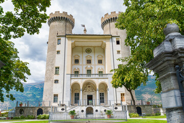The facade of the castle of the village named "Aymavilles", Aosta Valley, Italy, with towers at both sides. Dramatic clouds in the background. Green trees in the foreground. © Travelling Jack