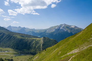 Fototapeta premium Sweeping panorama of lush green hills and rugged peaks under a bright blue sky, with soft clouds drifting above a sunlit alpine landscape in summer.