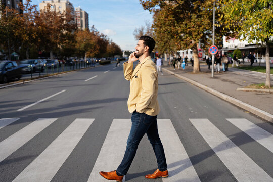 A stylishly dressed man in a tan blazer uses his smartphone while walking across a zebra crossing on an urban road during autumn.