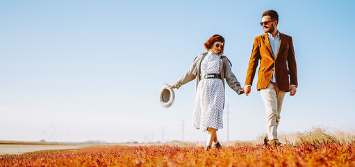 A couple stands together in a field overgrown with bright orange and yellow plants. They smile and embrace, enjoying the warm autumn sun. The photo features a clear sky and an open landscape. © maxbelchenko