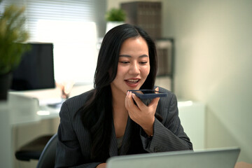 Obraz premium Professional woman using voice assistant chatbot voice on smartphone while working on laptop in modern office