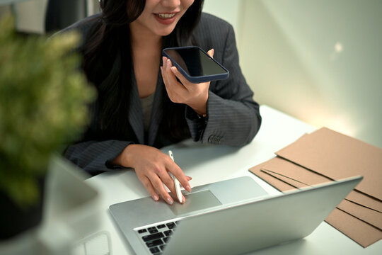 Professional woman using voice assistant chatbot voice on smartphone while working on laptop in modern office