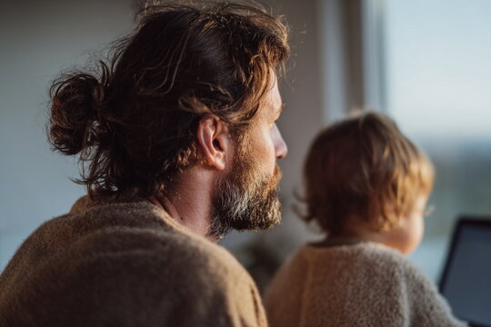 Side view of a father with his young child sitting by a window, watching a laptop. Soft daylight creates an intimate atmosphere of learning, curiosity, and connection.
