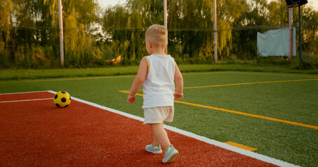 Obraz premium A cheerful little boy walks towards a ball on the tennis court. The sun sets in the background, creating a warm and joyful atmosphere during playtime.