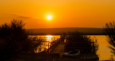 Silhouettes of individuals stroll towards a pontoon on a beautiful lakeside during sunset, creating a serene and picturesque evening atmosphere in nature. © RecCameraStock