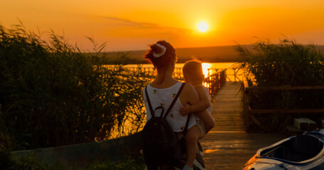 A silhouette of a mother carries her child while walking on a pontoon towards a serene lake during a beautiful sunset, surrounded by nature's tranquility. © RecCameraStock
