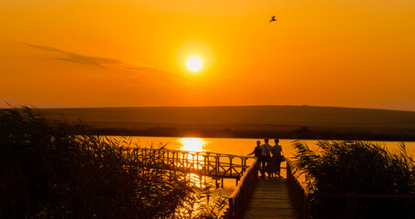 Silhouettes of individuals capture the beauty of a vibrant sunset on a tranquil lake from a pontoon. Nature's colors create a stunning backdrop for cherished memories. © RecCameraStock