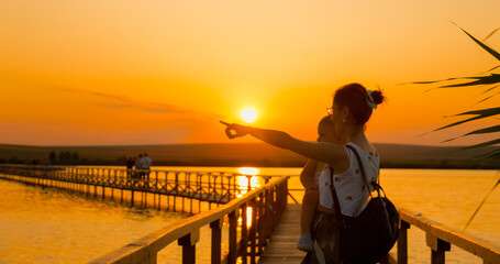 A mother carries her child while walking along a wooden pontoon at sunset. The tranquil scene highlights the beauty of nature and the bond between parent and child. © RecCameraStock