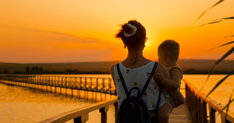 A mother carries her child while walking along a wooden pontoon during a stunning sunset. The sky glows with warm colors as they enjoy nature's beauty together. © RecCameraStock