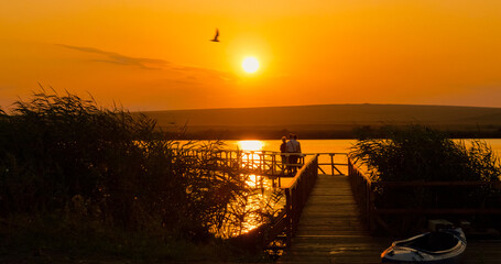 People gather on a pontoon at sunset, taking pictures against the backdrop of a beautiful lake and tranquil nature. The sky glows with warm colors as the day ends. © RecCameraStock
