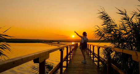 A mother strolls along a wooden pontoon, cradling her child in her arms as the sun sets behind them, casting warm colors across the serene waters. © RecCameraStock