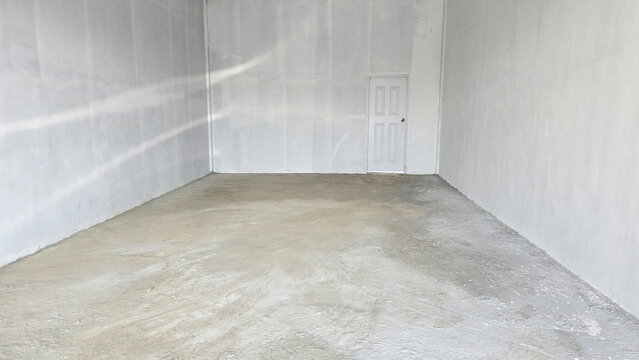The unfinished room area has white-painted door on the right. Inside, floor is bare concrete and walls are painted with white primer. Empty space inside building features rough-finish concrete floor .