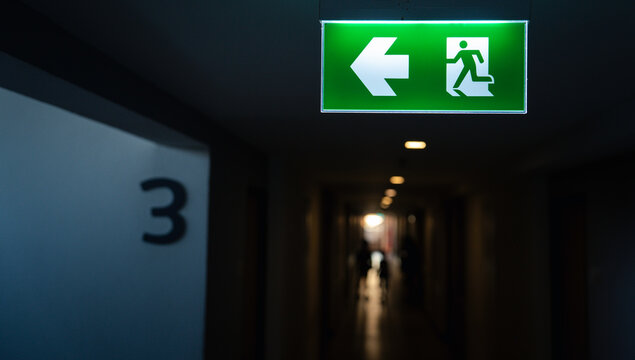 Emergency exit sign with running man icon and directional arrow in dark corridor. Safety evacuation route guidance fire escape symbol building security and risk management concept.