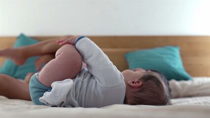 Baby lying on bed holding feet while laughing, capturing joyful and playful moments of early childhood in a relaxed and warm family environment