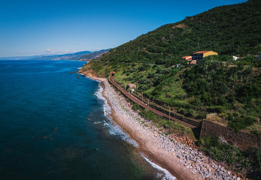 Aerial view of a coastal railway snaking along the rocky shore, a tunnel piercing the green hillside above the turquoise sea, Finale, Sicilia, Italy.