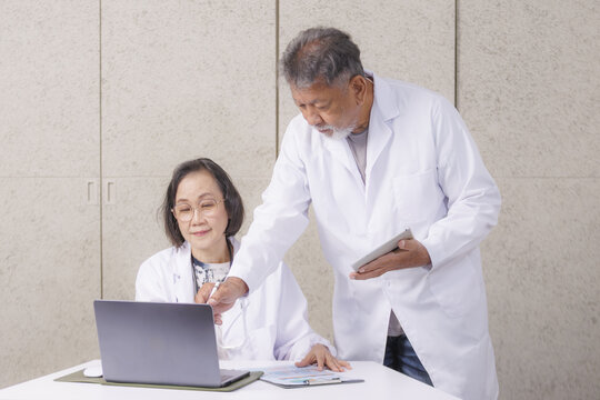 Elderly Asian doctors in white coats collaborate in medical office, with one using laptop and other holding tablet. They appear focused and engaged in their work