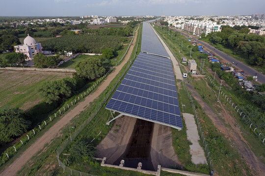 Aerial view of a solar panel covered canal cutting through green fields and urban development, contrasting earthy tones with the blue panels, Vadodara, Gujarat, India.