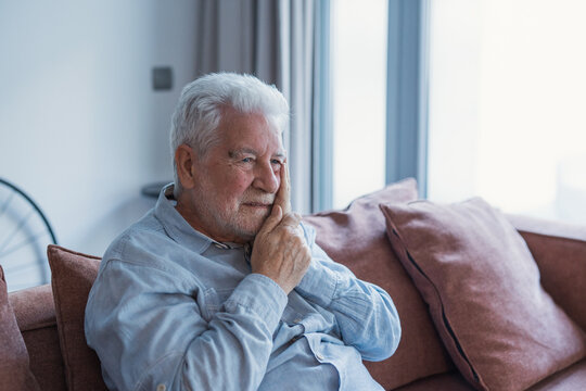 A senior man rests on the couch at home touching his jaw with a pained expression, reflecting dental pain and health concerns that disrupt his comfort and daily routine