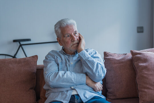 A lonely senior man sits on a sofa indoors staring outside, expressing concern, stress, and quiet despair while facing challenges and emotional strain in a silent home environment