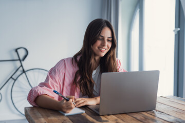 A focused woman sits at the table at home writing down useful information found online, organizing phone numbers, addresses, and daily tasks while managing household responsibilities © Daniel