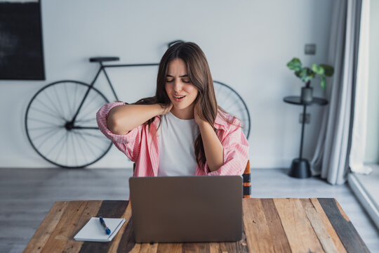A tired woman sits at a table rubbing her neck and tense shoulder muscles, suffering from chronic back pain caused by long laptop use, poor posture, and sedentary work affecting her health and comfort