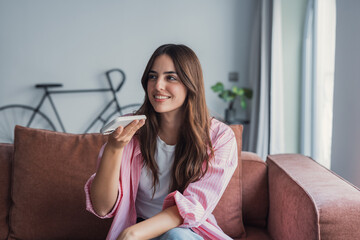 A happy young woman relaxes on a comfortable sofa at home, sending an audio message and sharing...