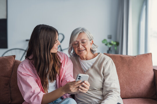 Smiling older mother and adult daughter using phone together, sitting on cozy sofa at home, happy young woman and mature mum looking at smartphone screen, watching video, having fun, two generations