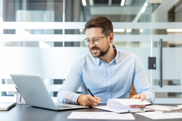 Obraz premium Young bearded businessman in glasses at a bright modern desk, focused on laptop and paperwork while writing notes confident, productive professional planning reports and analyzing data