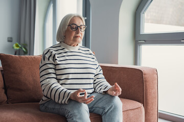 Obraz premium Mindful healthy mature woman practicing meditation at home, calm senior middle aged lady sitting on couch in lotus pose doing yoga for mental balance breathing air relaxing on stress free weekend
