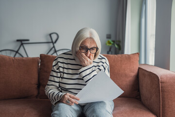 Fototapeta premium Upset elderly woman sitting on couch at home, reading document and getting stressful bad news at home. Frustrated senior lady shocked with letter, touching head, trying to cope with negative emotions