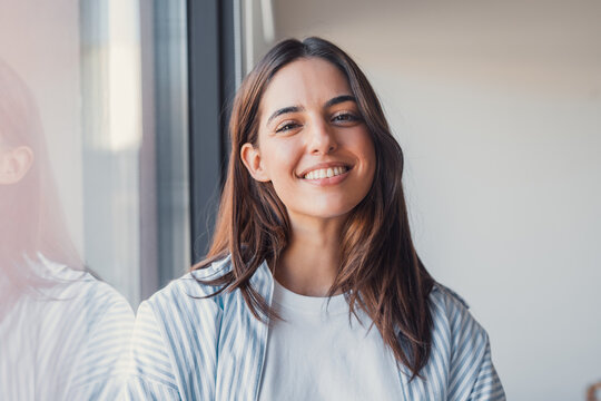 Portrait headshot of a confident, positive woman smiling naturally at the camera near a window, happy successful businesswoman or student posing for a professional profile photo