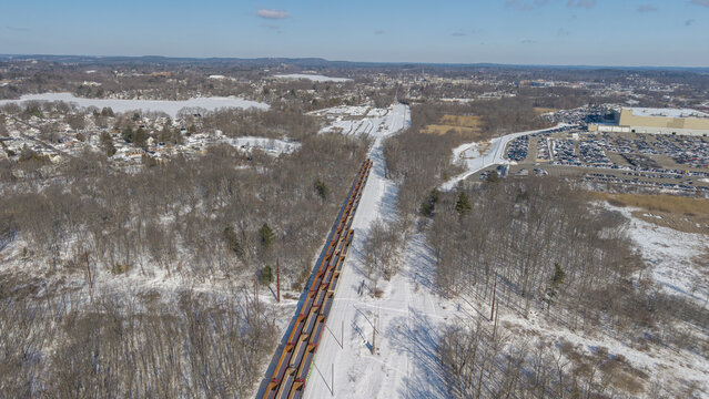 Aerial view of a long freight train cuts through a snowy landscape, with bare trees casting shadows on the pristine ground, Framingham, Massachusetts, United States.