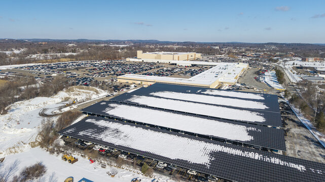 Aerial view of snow-dusted solar panels contrasting against a sprawling shopping mall and packed parking lots under a clear winter sky, Framingham, Massachusetts, United States.