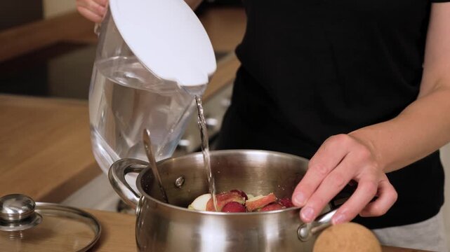 Pouring water into pot with fruits preparing homemade compote. Filling saucepan with liquid covering apples and plums for traditional drink. Adding clear water mixing base for sweet homemade beverage