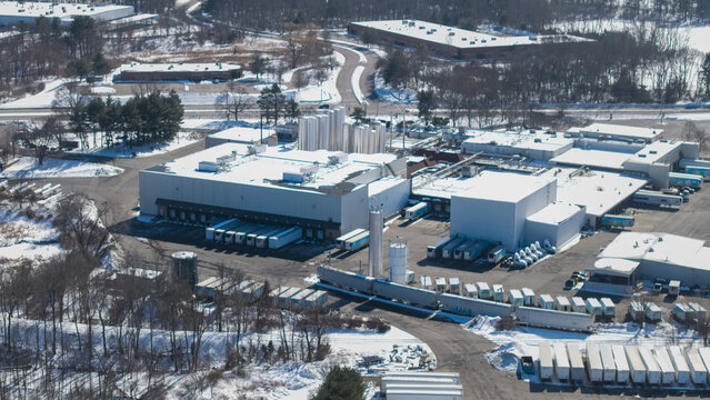 Aerial view of a sprawling industrial complex with boxy buildings and rows of parked trucks, nestled in a snow-dusted landscape, Bellingham, Massachusetts, United States.