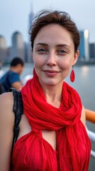 Fototapeta premium A woman wearing a red scarf and earrings is smiling at the camera. She is standing on a pier with a city skyline in the background
