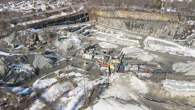 Aerial view of a stark, gray quarry carved into the earth, machines standing idle amidst the rocky landscape, dusted with the white of winter, Cumberland, Rhode Island, United States.