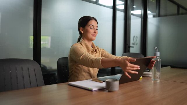 Young professional woman in beige shirt placing closed laptop into black bag after work at wooden desk in modern office