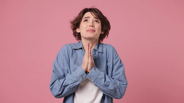 Pleading young woman she wears blue shirt white t-shirt casual clothes hold hands folded in prayer begging about something making wish isolated on plain pastel light pink background. Lifestyle concept
