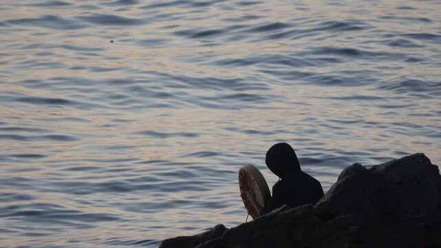 Drummer ritual shore plays a shamanic drum at sunset on the rocky coastline by the ocean waves, focusing on solitude.