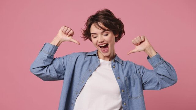 Confident happy young woman she wears blue shirt white t-shirt casual clothes pointing fingers on herself blink wink eye showing thumb up like gesture isolated on plain pastel light pink background