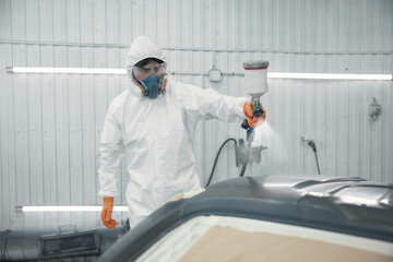 Automotive technician spray-painting car panel in protective suit inside paint booth