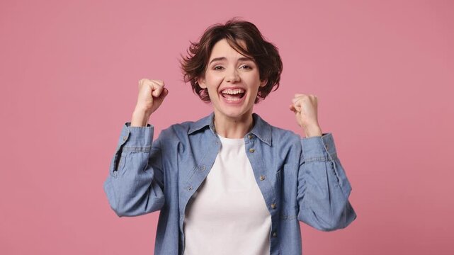 Excited jubilant overjoyed happy young woman she wearing blue shirt white t-shirt casual clothes doing winner gesture celebrate clenching fists say yes isolated on plain pastel light pink background
