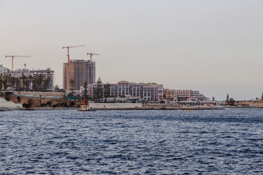 View of the skyline where modern buildings under construction contrast with established hotels, reflecting the soft light on the water, St. Julian's, Malta.