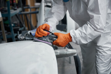 Auto body technician in a protective suit meticulously sands a car hood at a repair shop