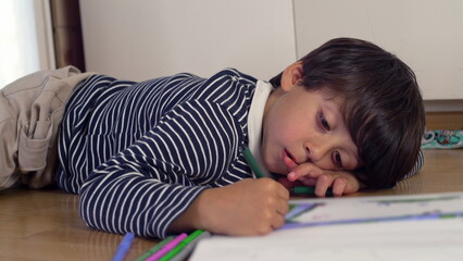 Young boy lying on the floor, drawing with a green marker, appearing relaxed yet focused, deeply engaged in creative expression, imagination, and artistic learning at home