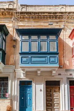 View of a vibrant blue enclosed wooden balcony and colorful doors adorn the aged facades of buildings bathed in sunlight, Rabat, Malta.