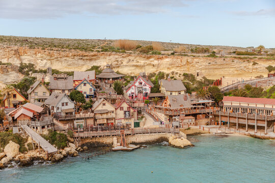 View of vibrant, quirky buildings nestled against rocky cliffs meet the turquoise sea in a picturesque coastal village, Il-Mellieha, Malta.