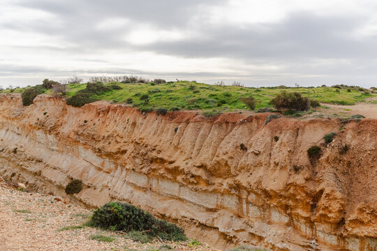 View of stratified earth cliff face, crowned with vibrant green vegetation under a cloudy sky, creating a striking contrast in Il-Mellieha, Malta.