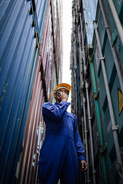 Logistics worker communicating by radio while inspecting shipping containers in a port yard, ensuring safety, coordination, and efficient cargo operations in global supply chain.
